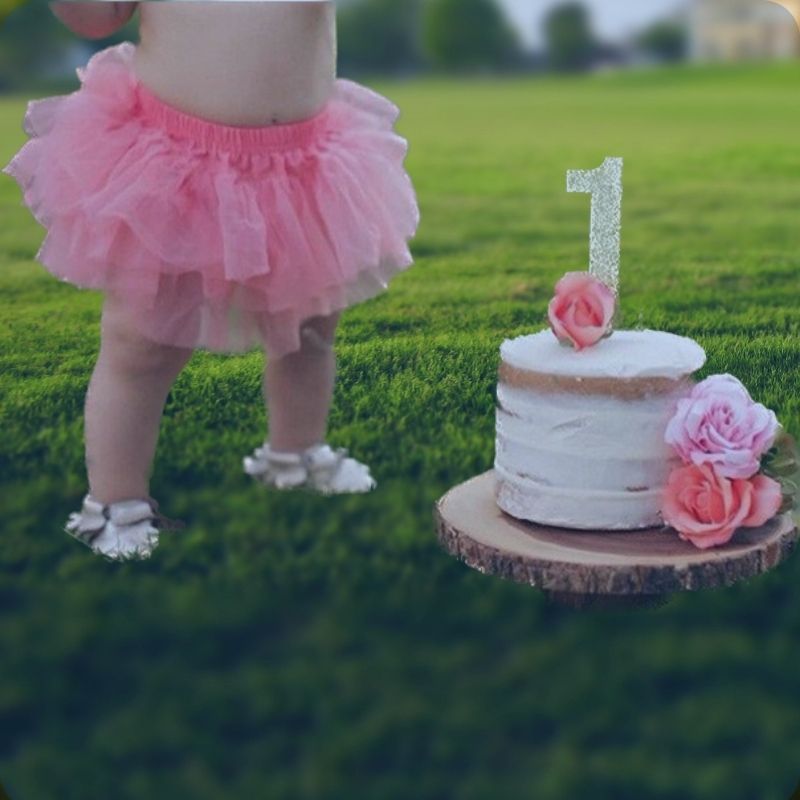 Adorable baby girl wearing Dear Pastel's Tutu Skirt, standing next to her birthday cake with a '1' candle. A sweet and joyous moment captured on a lush grass lawn.